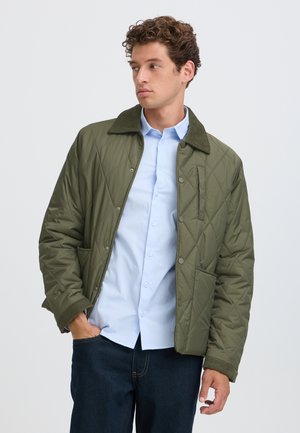 Un homme porte une veste matelassée vert olive sur une chemise bleu clair et un jean foncé. Il se tient debout, une main dans la poche.
