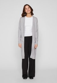 Heledhalli pikk kardigan ribadega servades ja kahe ees oleva taskuga, kantud valge topi ja mustade laia säärega pükste peal, neutraalsel taustal.