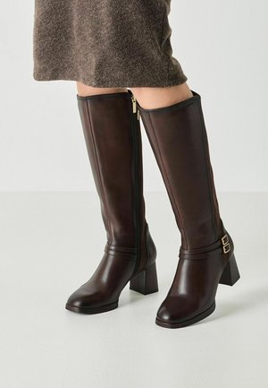 Femme portant des bottes en cuir marron foncé montant jusqu'aux genoux avec des talons blocs et des fermetures éclair latérales, associées à une jupe en laine marron.