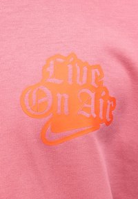 Pomarańczowy napis "Live On Air" z logo Nike Swoosh na różowym, teksturowanym tle tkaniny.