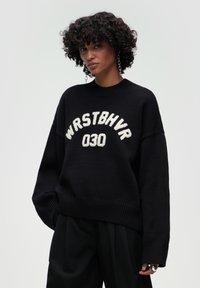 Czarny sweter z dzianiny z białym napisem "WRSTBHV 030." Oversized krój, dekolt typu crew neck oraz ściągacze na rękawach i u dołu. Teksturowany materiał.