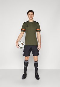 Groen sportshirt met oranje accenten, zwarte shorts, zwarte sokken met witte strepen en zwarte voetbalschoenen. Houdt een voetbal vast.