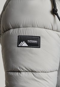 Chaqueta acolchada gris claro con patrón de cuadrícula, dobladillos elásticos y una parcha negra con logotipo en blanco. Acabado texturizado.