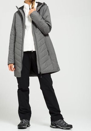 Personne portant une veste matelassée grise à capuche, un pull zippé blanc, un pantalon noir et des chaussures de randonnée noires, se tenant sur un fond neutre.