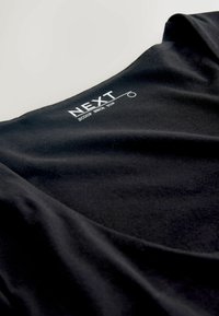 Melnā blūze ar apaļu kakla izgriezumu, izgatavota no mīksta auduma, ar gludu tekstūru un zīmola nosaukumu "NEXT" drukātu baltā krāsā kakla izgriezumā.