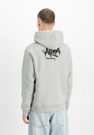 Mann trägt hellgrauen Kapuzenpullover mit schwarzem "Alpha Industries"-Logo am oberen Rücken, kombiniert mit hellblauen Jeans, schaut weg.