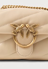Borsa a mano in pelle beige con design trapuntato, hardware dorato a forma di uccello, dettaglio con anello ovale e cuciture visibili. Nome del marchio "PINKO" impresso.