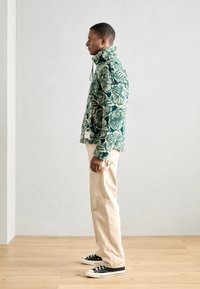 Giacca in pile verde e crema con motivo, colletto alto, abbinata a pantaloni beige e sneaker nere. Vista laterale, in posa in piedi.
