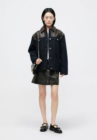 Veste en denim avec un col en cuir marron, tissu bleu foncé, deux poches avant, associée à une jupe en cuir noire, des mocassins noirs et un sac bandoulière.