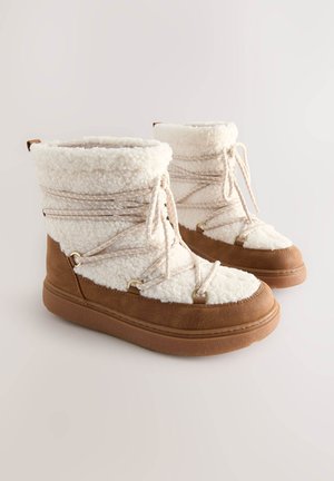 Cizme maro până la gleznă cu partea superioară din fleece alb pufos, prevăzute cu șireturi decorative și accesorii aurii. Talpile groase din cauciuc oferă aderență.