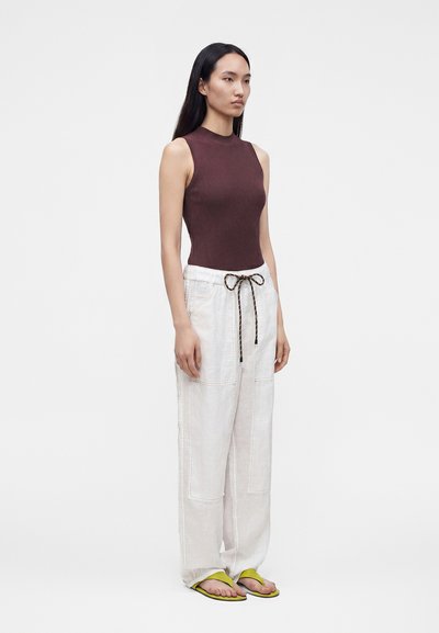 Femme debout portant un haut marron sans manches, un pantalon blanc ample avec taille à cordon et des tongs jaunes sur un fond uni.