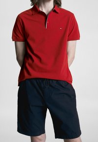 Czerwona koszulka polo z kołnierzykiem, krótkimi rękawami i małym logo na piersi, w parze z ciemnoniebieskimi szortami z elastycznym pasem.