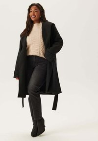 Donna che indossa un maglione a trecce color crema, jeans neri a vita alta, un cappotto nero appoggiato sulle spalle e stivali neri alla caviglia, sorridendo e camminando.