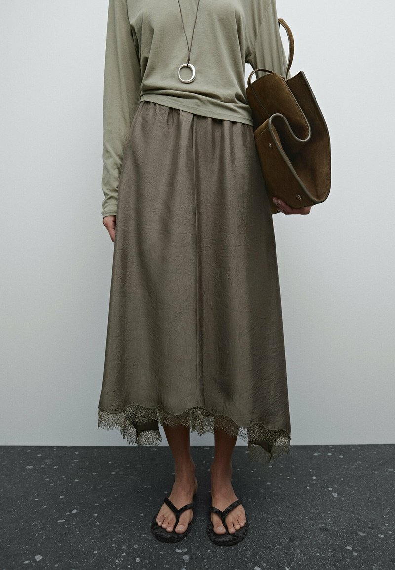 Personne portant une longue jupe olive avec ourlet en dentelle, un haut olive assorti, des tongs noires, un collier avec pendentif rond, et tenant un sac en cuir marron plié.