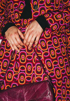 Patterned fabric coat with bold red, pink, orange, and purple circular designs; hands holding a shiny purple handbag, showing intricate detail.