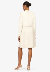 Hvid cropped jakke med blondebagdetalje parret med en struktureret hvid nederdel. Modellen bærer strappy hvide hæle, der viser en glat stoffinish.