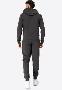 Mørkegrå hættetrøje og matchende joggingbukser, lavet af blødt materiale. Har en hætte, ribbede manchetter og elastisk talje. Hvide sneakers.
