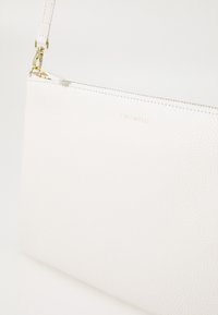 Pochette en cuir blanc avec une surface texturée, dotée d'une fermeture éclair dorée, d'un logo en relief à l'avant et d'une bandoulière ajustable.