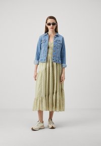 Chaqueta de mezclilla, azul clara, abotonada con bolsillos; vestido maxi verde con flores, con volantes, hasta el tobillo. Zapatillas deportivas blancas con acentos.