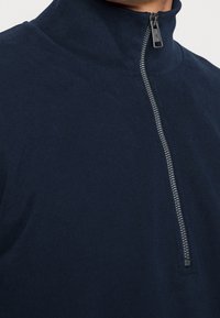 Pullover blu navy a collo alto con zip, realizzato in tessuto liscio e dotato di una cerniera metallica con tag pull rettangolare.