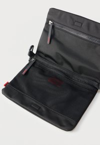 Μαύρο υφασμάτινο τσαντάκι με δύο θήκες με φερμουάρ, μπροστινή τσέπη από δίχτυ και κόκκινο λογότυπο "HUGO" στις τσουλήθρες των φερμουάρ και στην ετικέτα.