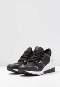 Svarta och vita träningsskor med ett strukturerat svart material, snörning och en slank vit mellansula med en subtil logotyp.