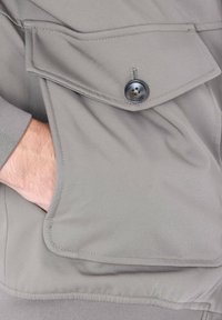 Giacca grigia chiara con una grande tasca frontale con chiusura a bottone, texture morbida e maniche con polsini.