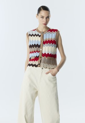 Donna che indossa un gilet in maglia senza maniche con motivo ondulato multicolore e bottoni, abbinato a pantaloni trapuntati crema a vita alta, mano in tasca.