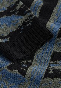 Close-up of knitted fabric with abstract black, blue, and gray patterns and a ribbed black cuff on the sleeve.