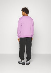 Fialová mikina s žebrovanými manžetami, v kombinaci s černými tepláky. Modelka je viděna zezadu, což ukazuje na volný střih a měkkou látku.