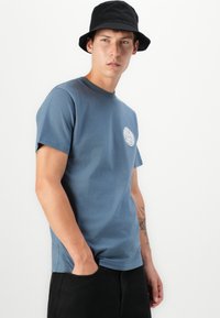 Blå bomull T-skjorte med rund hals og korte ermer, med et sirkulært grafisk logo. Båret med svarte shorts og en svart bøttehatt.