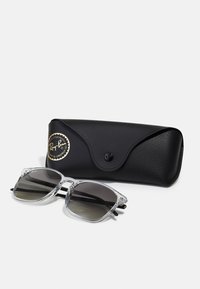 Ray-Ban solbriller med klar acetatramme og grå gradientlinser, ledsaget af et sort læderetui med et guld logo.