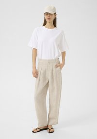 White short-sleeve t-shirt, beige high-waisted linen pants, and black flip-flops. Light-colored baseball cap and simple earrings.