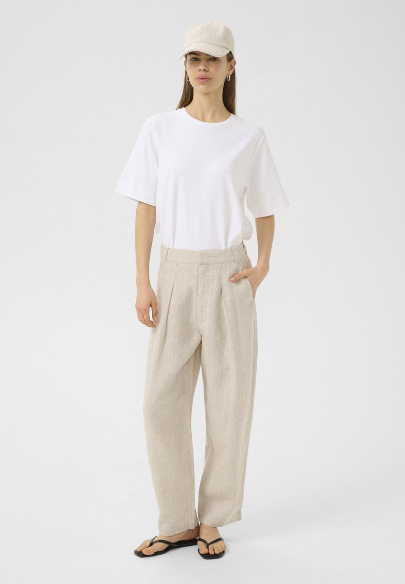 White short-sleeve t-shirt, beige high-waisted linen pants, and black flip-flops. Light-colored baseball cap and simple earrings.