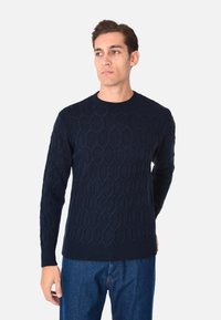 Maglione lavorato a maglia blu navy con un motivo esagonale testurizzato, scollo rotondo e maniche lunghe, abbinato a jeans di denim.