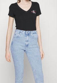 Camiseta de algodón negra con cuello en V, que presenta un logotipo de CK en blanco y rojo, combinada con unos jeans de talle alto de color azul claro. Textura suave, diseño ajustado.