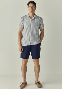 Kortærmet skjorte med knaplukning i blå og hvide lodrette striber, parret med marineblå shorts. Brune strukturerede sko fuldender outfittet.