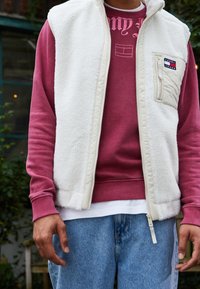 Biała kamizelka z polaru na suwak noszona na bordowej bluzie, z naszytą kieszenią z logo na piersi. Do tego jasnoniebieskie dżinsy.