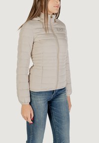 Giacca imbottita beige con colletto alto, chiusura con zip e polsini elasticizzati. Presenta un logo discreto e un design a taglio slim.