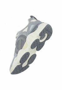 Zapatilla deportiva gris con materiales de malla y gamuza, forma redondeada, suela blanca gruesa y suela exterior con patrón en zigzag. Presenta la marca en la lengüeta.