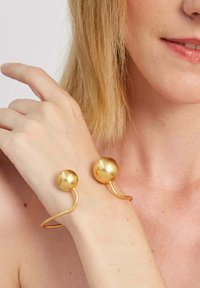 Zlatý kovový náramok s dvoma guľatými guľami na oboch koncoch, s hladkým, odrážajúcim povrchom a zakriveným, otvoreným dizajnom.