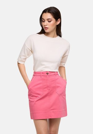 Mujer con cabello liso y castaño, lleva una blusa blanca de manga corta y una falda rosa de talle alto con bolsillos delanteros, con las manos metidas en los bolsillos.