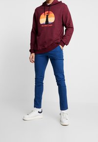 Bordová mikina s kapucí a grafickým potiskem, zobrazující západ slunce u majáku s textem "Severní mys". Spárováno s modrými slim-fit kalhotami a bílými teniskami.