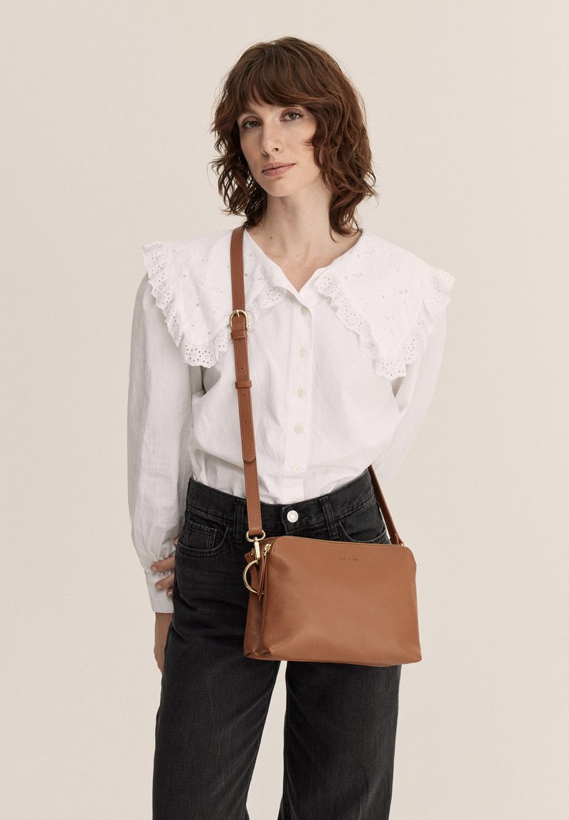Brun læder crossbody taske med tekstureret overflade, gulddetaljer og slankrem. Modellen bærer en hvid bluse med hulmønster og sorte jeans.