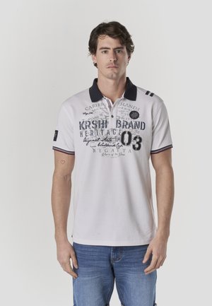 Polo blanco con cuello azul marino, con texto impreso y detalles de logotipo. Mangas cortas con detalle de rayas en los puños. Combinado con vaqueros azules.