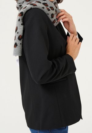 Vrouw die een grijze sjaal met luipaardprint om haar hals legt, gekleed in een zwarte blazer en blauwe spijkerbroek, tegen een effen achtergrond.