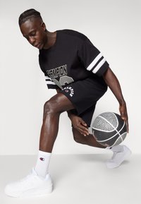 T-shirt gráfica preta da Champion com riscas brancas, calções pretos, sapatilhas brancas da Champion, e meias brancas, segurando uma bola de basquete texturizada.