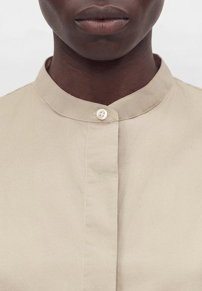 Το κάτω μέρος του προσώπου και ο λαιμός ενός ατόμου με σκουρόχρωμο δέρμα που φορούν μια μπεζ πουκάμισα με γιακά τύπου μανταρίνι, κλεισμένο με ένα λευκό κουμπί.