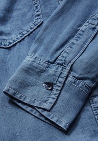 Denimskjortärm i ljusblått, med knappstängning och sydda detaljer. Slät textur med synlig vävstruktur och subtil blekning.