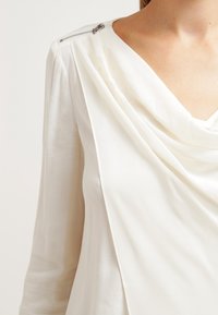 Weiße, drapierte Bluse mit glatter Textur, verziert mit einem silbernen Reißverschlussakzent an der Schulter und einem tiefen V-Ausschnitt.